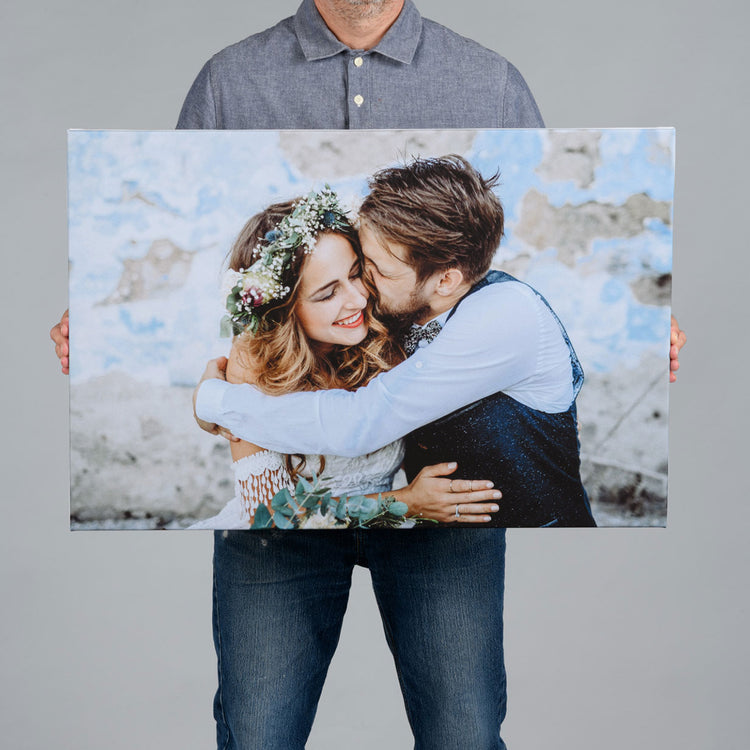 A man holds up a 75X50cm canvas print showing a smiling wedding couple embracing, against a plain light gray studio background.