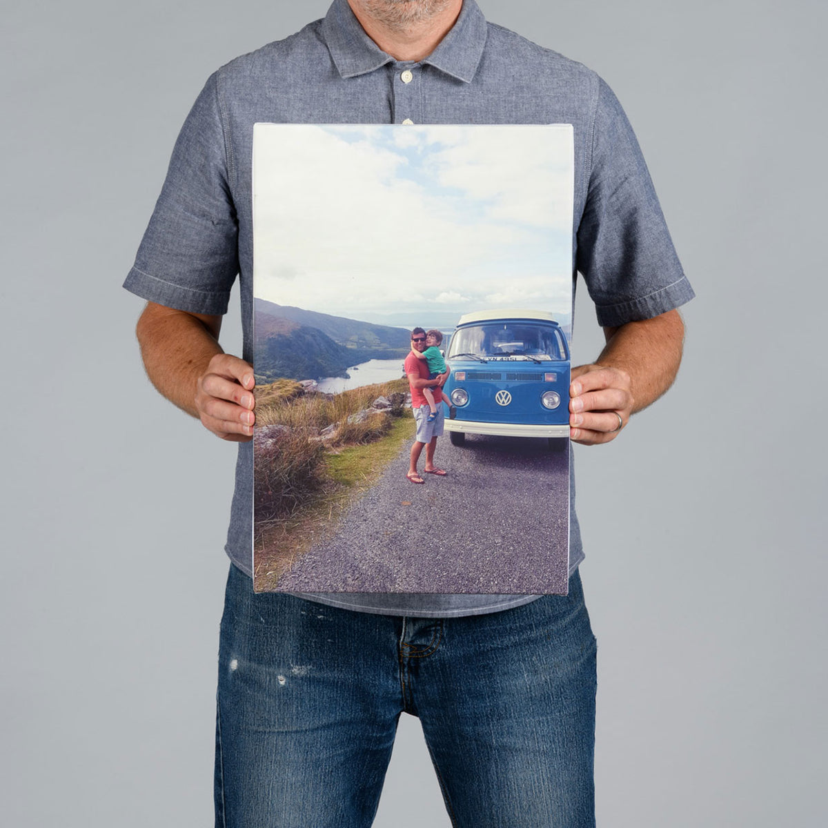 A man from The Canvas Works holds up a 30x45cm rectangular photo canvas print showing a blue vintage Volkswagen bus on a road with mountains and water in the background.