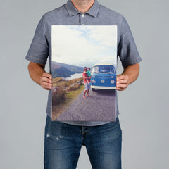 A man from The Canvas Works holds up a 30x45cm rectangular photo canvas print showing a blue vintage Volkswagen bus on a road with mountains and water in the background.