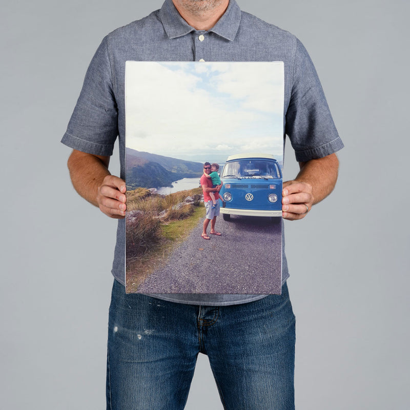 A man from The Canvas Works holds up a 30x45cm rectangular photo canvas print showing a blue vintage Volkswagen bus on a road with mountains and water in the background.