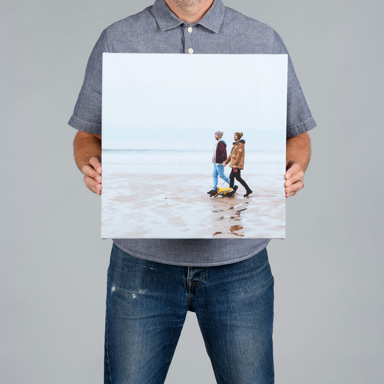 A man from The Canvas Works  holds up a square 40x40cm canvas print showing a beach scene with two people walking on the sand.