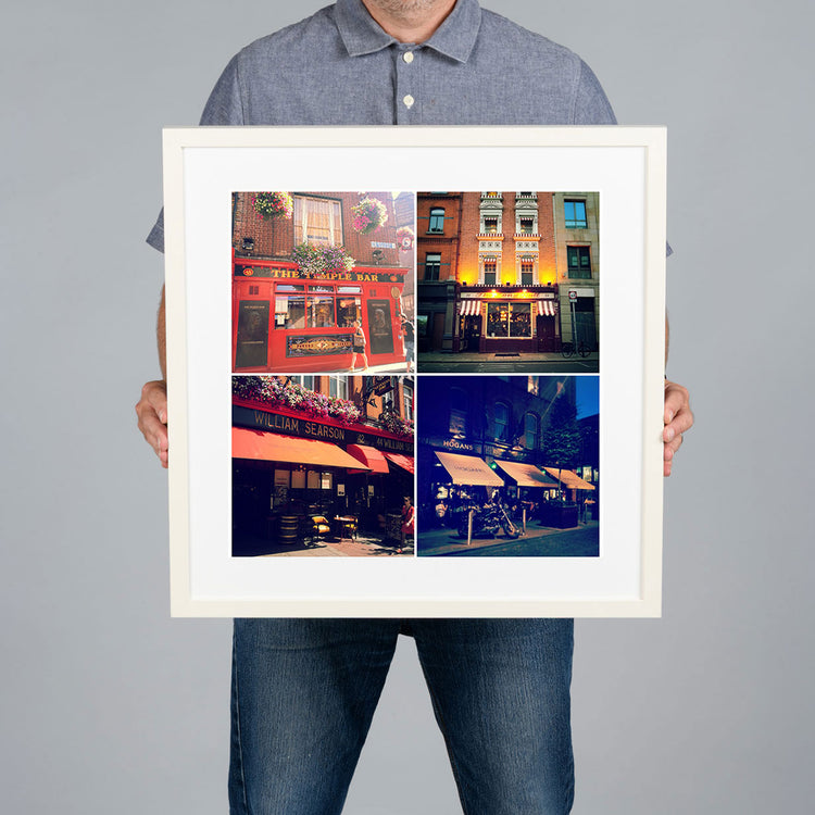 A man holds a white-framed photo print with a white mat. The print features a 2x2 collage photo grid of famous Dublin pubs. The print is 40x40cm and the frame is 54x54cm.  