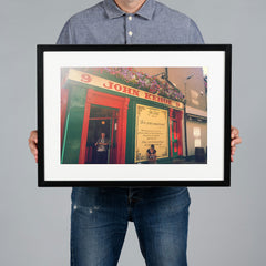 A person holding a 45x30cm white-framed art print of a colorful pub exterior with red and green facade details, set against a solid gray background. The pub is Kehoes in Dublin. 