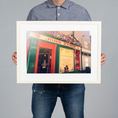 A person holding a 45x30cm white-framed art print of a colorful pub exterior with red and green facade details, set against a solid gray background. The pub is Kehoes in Dublin. 