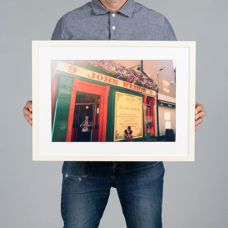 A person holding a 45x30cm white-framed art print of a colorful pub exterior with red and green facade details, set against a solid gray background. The pub is Kehoes in Dublin. 