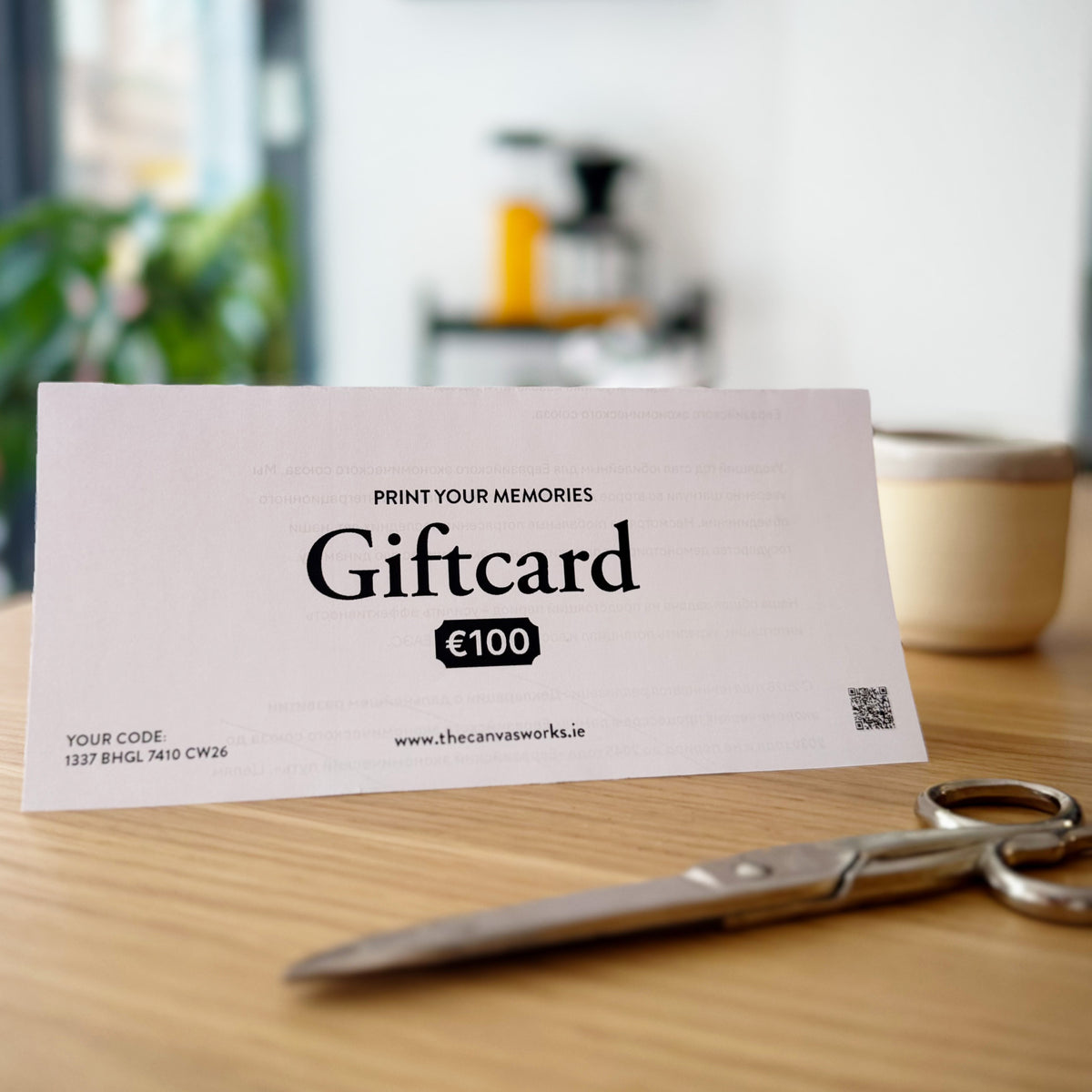 A printed gift card from The Canvas Works with '€100' and the text 'Print your memories Giftcard' sits on a wooden tabletop. Scissors are in the foreground and a blurred kitchen or cafe setting is visible in the background.